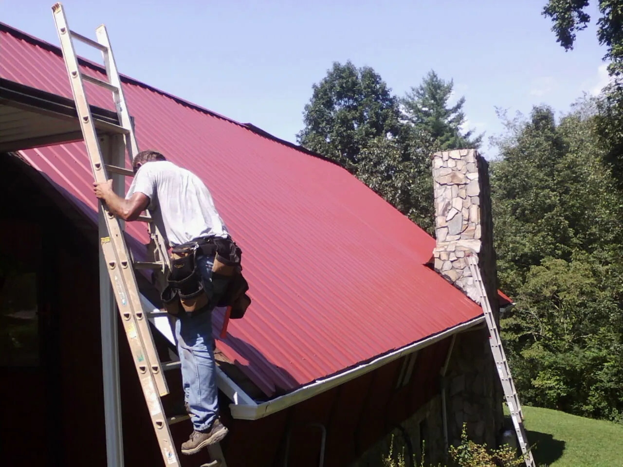 Roofer on a ladder beside a red metal roof and stone chimney — Soffit & Fascia Repair by Roofing Brothers in Wamego